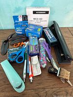 Various office supplies arranged on tabletop including staplers, hole punch, pens, tapes, scissors, and boxes of staples
