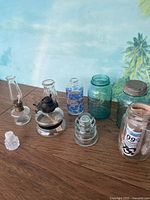 View of assorted glass items including 2 vintage hurricane lamps, small milk bottle, and glass insulator on wooden surface with tropical background