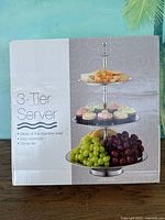 Box showing front image of the 3-tier stainless steel server with food items on each tier illustrating its use.