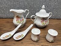 Porcelain creamer, sugar jar with lid, two spoon rests, and salt and pepper shakers arranged on wooden surface showing full set.