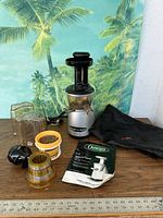 Omega VRT 350 fruit and vegetable juicer with accessories on table including pulp container, juice container, two strainers, black plunger, carrying bag and manual
