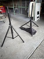 Two black tripod style adjustable speaker stands set up and standing on a concrete floor with a black carrying bag lying between them.