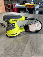 Side view of Ryobi electric orbital sander showing ergonomic handle, dust collection bag, and power cord.