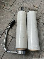 Photo showing two rolls of white stretch wrap and a chrome metal handle with a black grip resting on a paved surface.