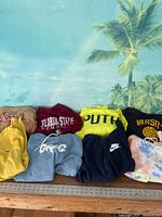 Overview of 8 assorted sweatshirts showing various colors and designs on a light blue background with palm trees.