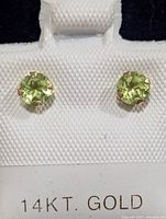 Close-up photo of the front of two round green peridot stud earrings set in 14KT gold prong mounting on a white display card.