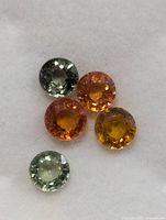 Five loose faceted round sapphires on white paper background, showing mixed yellow, orange, and green colors.