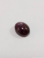 Single polished oval cabochon star ruby gemstone, purple-red color, showing asterism effect and natural inclusions.