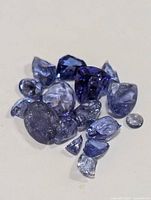 Close-up image showing a loose collection of tanzanite gemstones in a variety of shapes and sizes, including oval, pear, and round cuts, with blue and violet coloration.