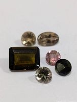 Photo showing six loose tourmaline gemstones in different cuts and colors including rectangular, oval, and round shapes in brown, pink, and green shades.