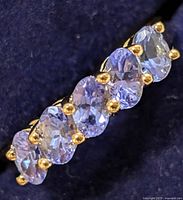 Close-up of silver ring with seven light purple oval tanzanite stones secured by gold prongs against dark background.