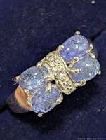 Close-up of silver ring showing four light purple blue tanzanite stones with prong setting and central silver section with small clear accent stones on dark blue background.