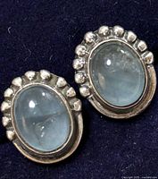 Pair of silver earrings with genuine gemstones shown on dark background. Each earring features an oval cabochon stone with a silver beaded border.