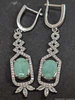 Pair of sterling silver earrings featuring oval green emeralds and cubic zirconia accents in a decorative pattern with hinged backs.