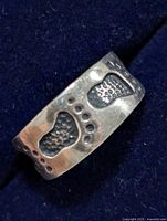 Close-up of silver toe ring showing a wide band with embossed footprint designs and circular decorative dots, set against a dark fabric background.