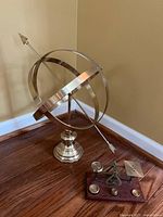 Full view of armillary sphere and scale side by side on wooden floor against a beige wall.