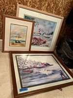Three framed waterfront landscape watercolor paintings arranged on floor, including beach scene, lighthouse, and coastal village.