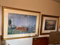 Photo of two framed nautical prints hung on wall showing boats docked at marina with calm water and sky. Gold frames, cream mats, glass covered.