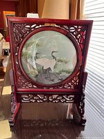 Front view shows detailed wooden frame with intricate carvings surrounding fabric artwork of cranes and pine trees, mounted on stand.