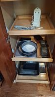 Items inside a wooden cabinet including the Hamilton Beach hand mixer on the top shelf and various bakeware pans including springform and rectangular pans on bottom shelves.