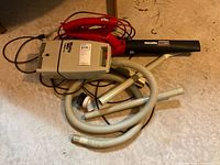 Photo showing red and black Homelite electric blower with power cord, beige Mighty Mini Shop Vac, flexible hose, and two metal extension tubes arranged on the floor.