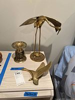 Wide view of three items on a light-colored cabinet: a tall brass bird sculpture, a brass duck wall hanging, and a brass ornate candle holder.