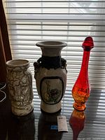 Photo of all three items: one red and amber glass decanter with stopper, two ceramic vases, one with face relief and crackle glaze, one with floral and fruit raised decorations.