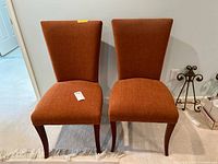 Front view of two matching dining chairs upholstered in orange-brown fabric, showing the seat and backrest details