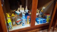 Photo of cabinet interior showing assorted cleaning spray bottles including Windex, Febreze, Mr. Clean, Scrub Free, Suave Scents, along with boxes of SOS pads and brushes.