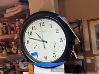 Round wall clock with white face and black frame showing time, temperature, and humidity.