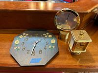 Painted slate octagon clock with floral design positioned on wooden shelf with two other clocks.