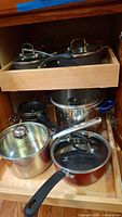 Cookware stored in wooden drawer including pots and pans of various sizes and materials, some with glass lids.