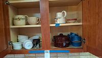 Cabinet interior showing various Corningware and ceramic pitchers, soup cup, mugs, and bowls on shelves