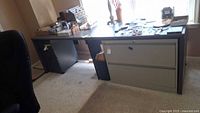 Long rectangular office desk with grey Formica top and two beige metal filing cabinets on either side. Desk is set up in a room with beige carpet.