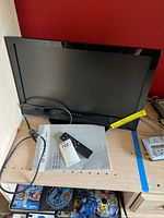 22-inch Dynex TV on wooden shelf with power cord, remote control, and user manual placed in front of the TV.