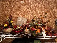 Shelf display of six faux floral arrangements with autumn colors, incorporating flowers, leaves, berries, and decorative bases including woven baskets and bowls.