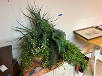 Terracotta planter filled with faux grasses, ferns, and leafy vines placed on a table near a framed picture.