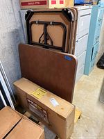 Three folding tables visible: two rectangular with brown laminate tops and folding metal legs, one square table with similar brown top and legs, placed in basement corner.