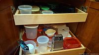 Kitchen cabinet drawer with a variety of plastic food storage containers, Pyrex glass bowls with lids, kitchen grater, measuring cups, and spoons, all needing cleaning.
