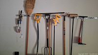 A wall-mounted rack displaying garden tools including two shovels with D-grip handles, a garden fork, a broom, a cutter, and a silver metal air pump. Tools are hanging evenly spaced on pegs.