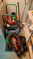 Overview of gardening tools and supplies arranged in basement corner including nozzles, soft flex boots, extension cord with light, metal hole maker, sickle, green garden cart, and orange bowl with small parts.