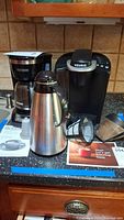 Photo showing Black & Decker 12-cup drip coffee maker, Keurig K-Classic coffee maker, stainless steel Gevalia carafe, coffee filter, and instruction manuals on countertop.