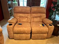 Front view of the tan suede-like La-Z-Boy power reclining loveseat with two reclining seats and black cup holders in the armrests. Remote controls are placed on each seat.