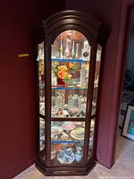 Front view of lighted wooden corner curio cabinet filled with various decorative glassware and ceramics, showing four adjustable blue glass shelves.