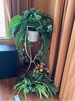 Full view of a white metal plant stand holding a white wicker basket filled with faux plants including ivy and ferns, with additional faux plants at the base.