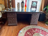 Front view of dark wood office desk with glass top, placed on a wooden floor with a patterned round rug underneath. Various decorative items are on top of the desk.