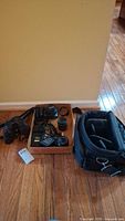 Overview photo of all lot items laid out on wood floor showing Olympus camera body, lenses, charger, flash, binoculars, and camera bag.