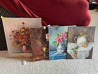 Four still life paintings leaning against a red chair; three on canvas, one on board, all featuring floral and vase subjects.