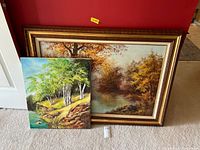 Photo displaying both oil paintings together, one framed with autumn foliage scene, one unframed with green birch tree scene, on carpeted floor with white door and red wall background.