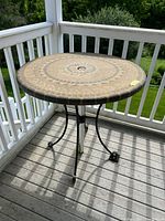 Full view of round patio table showing mosaic resin top with umbrella hole and three-legged metal base on outdoor deck with railing.